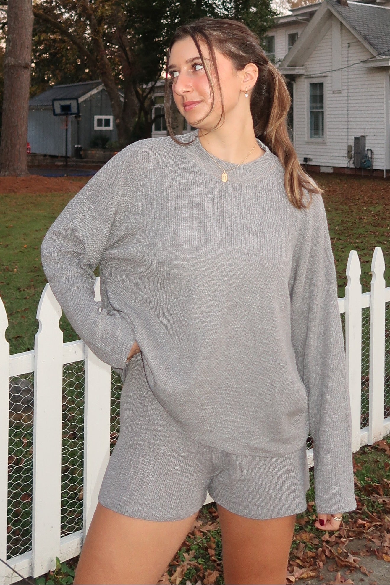 woman wearing a light gray oversized long sleeve top and matching shorts, standing outdoors by a white fence