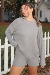 woman wearing a light gray oversized long sleeve top and matching shorts, standing outdoors by a white fence