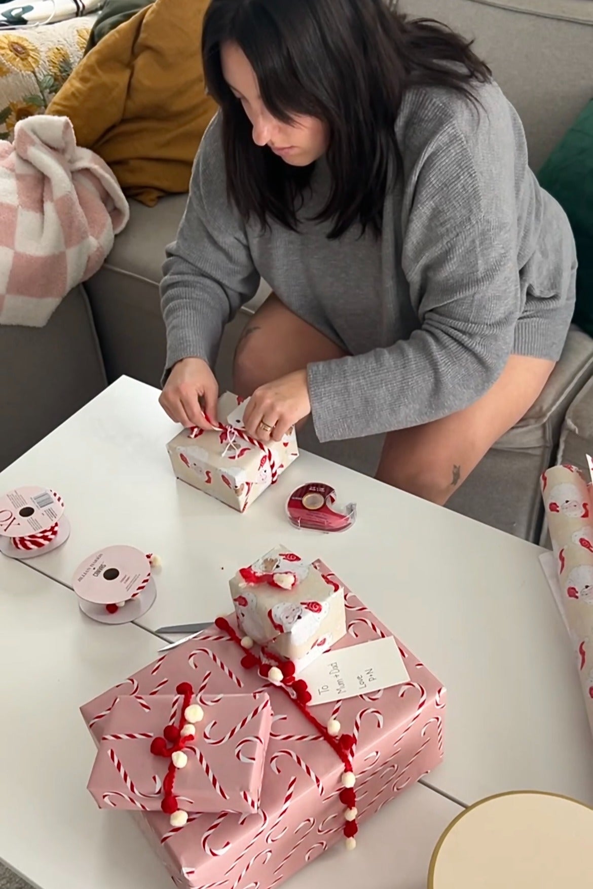 woman wearing a gray sweater sitting at a table wrapping holiday gifts with festive wrapping paper and ribbons