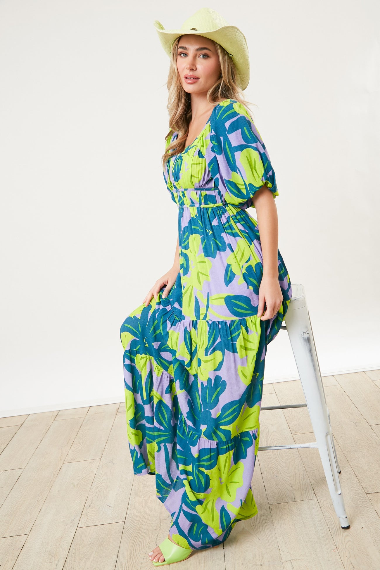 woman wearing a colorful floral maxi dress with puff sleeves and a wide-brimmed hat, sitting on a stool