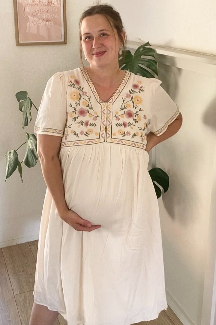 woman wearing a cream maternity dress with floral embroidery and holding her baby bump