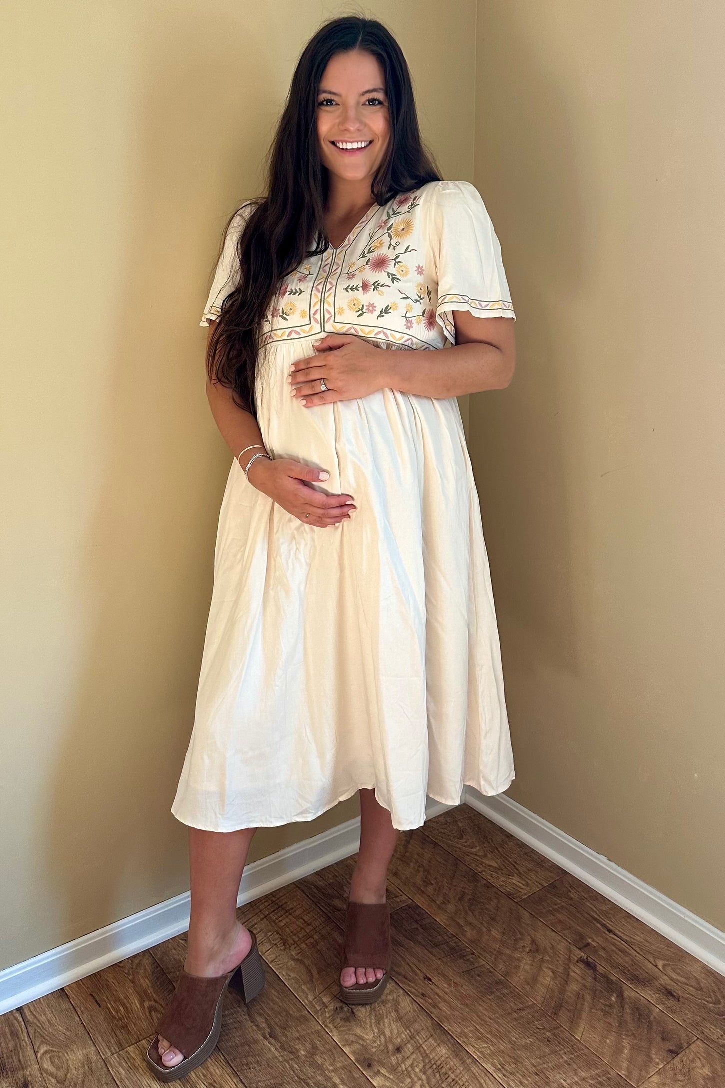 woman wearing a cream maternity dress with floral embroidery and holding her baby bump