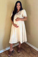 woman wearing a cream maternity dress with floral embroidery and holding her baby bump
