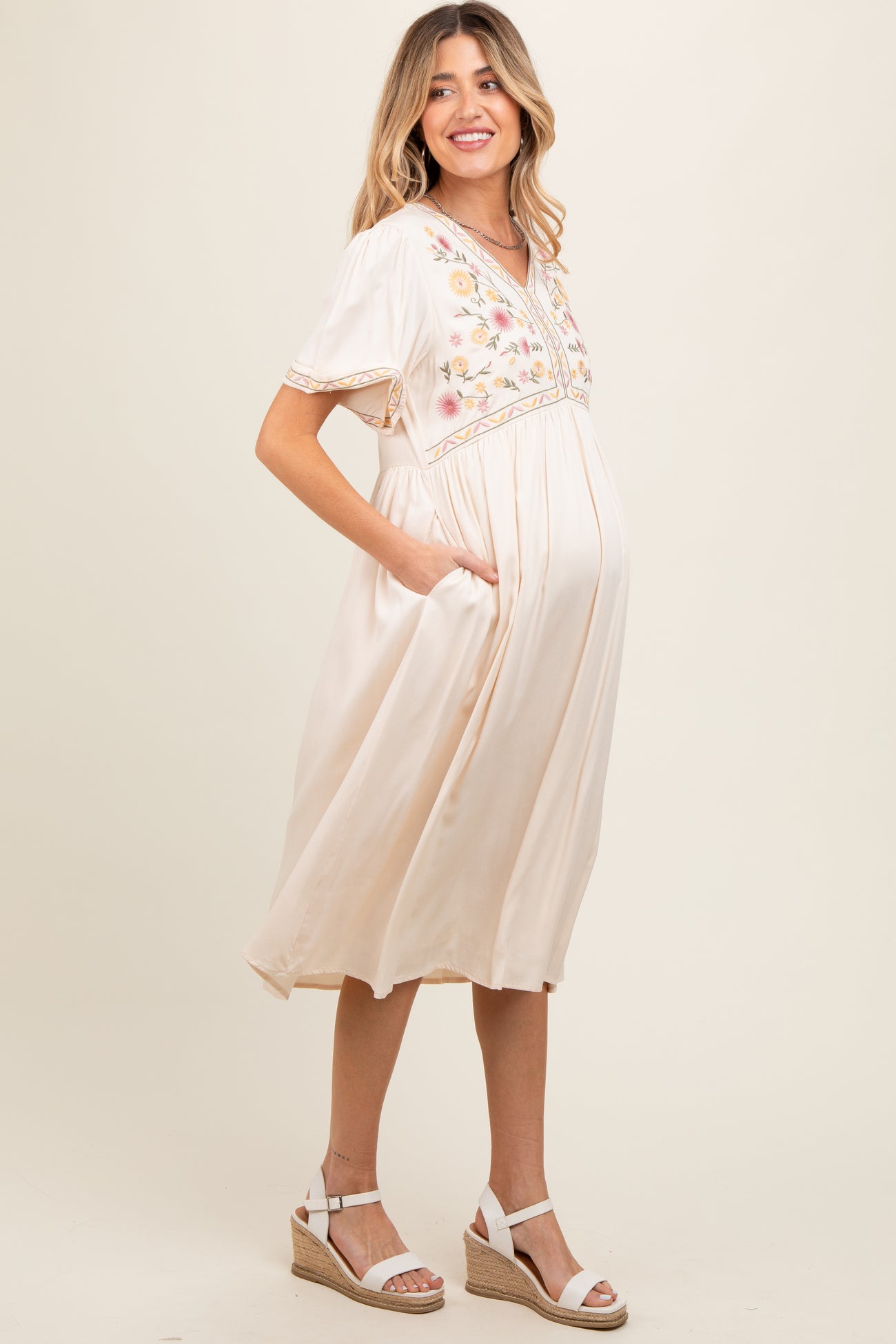 woman wearing a cream maternity dress with floral embroidery and pockets, showing her baby bump