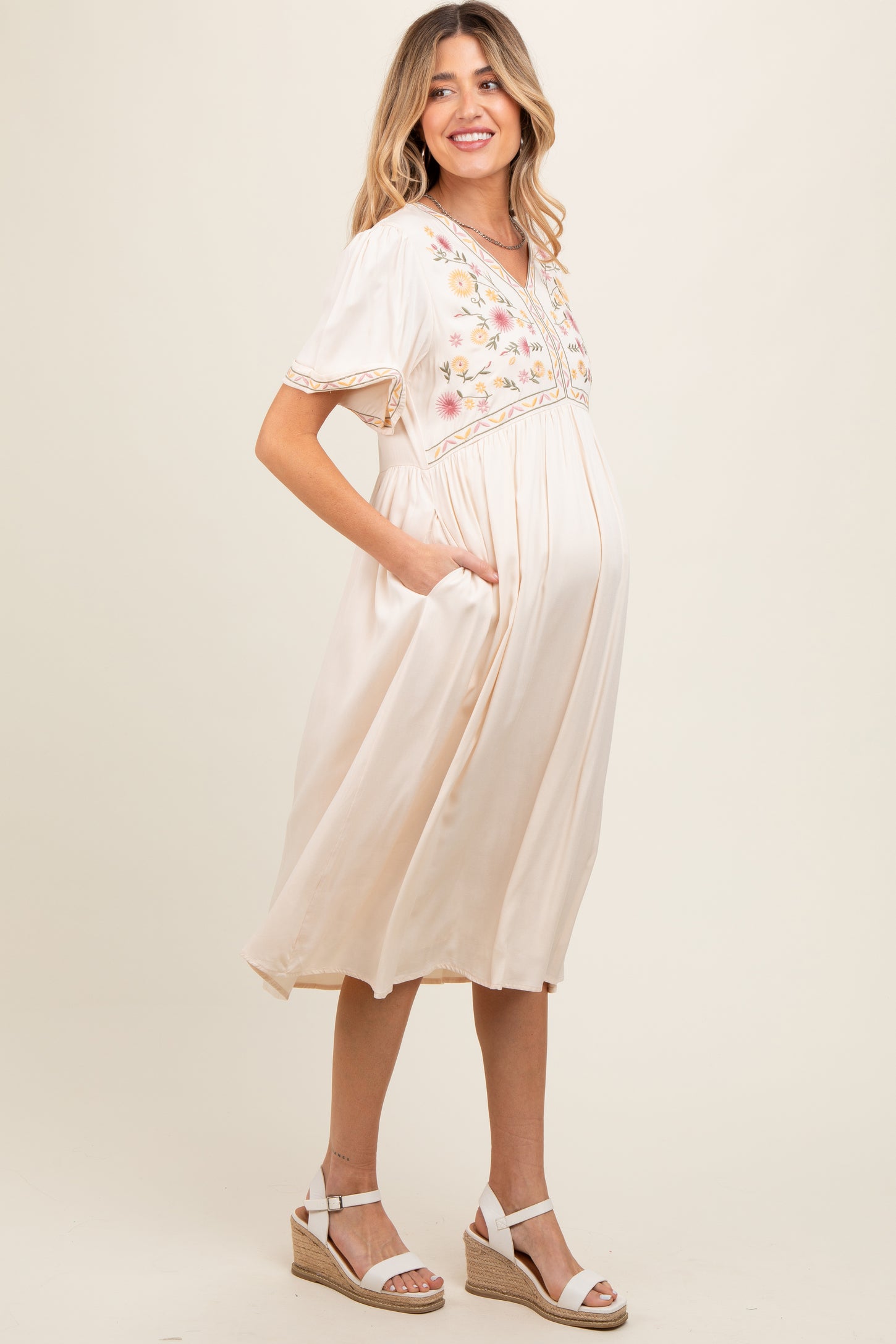 woman wearing a cream maternity dress with floral embroidery and pockets, showing her baby bump