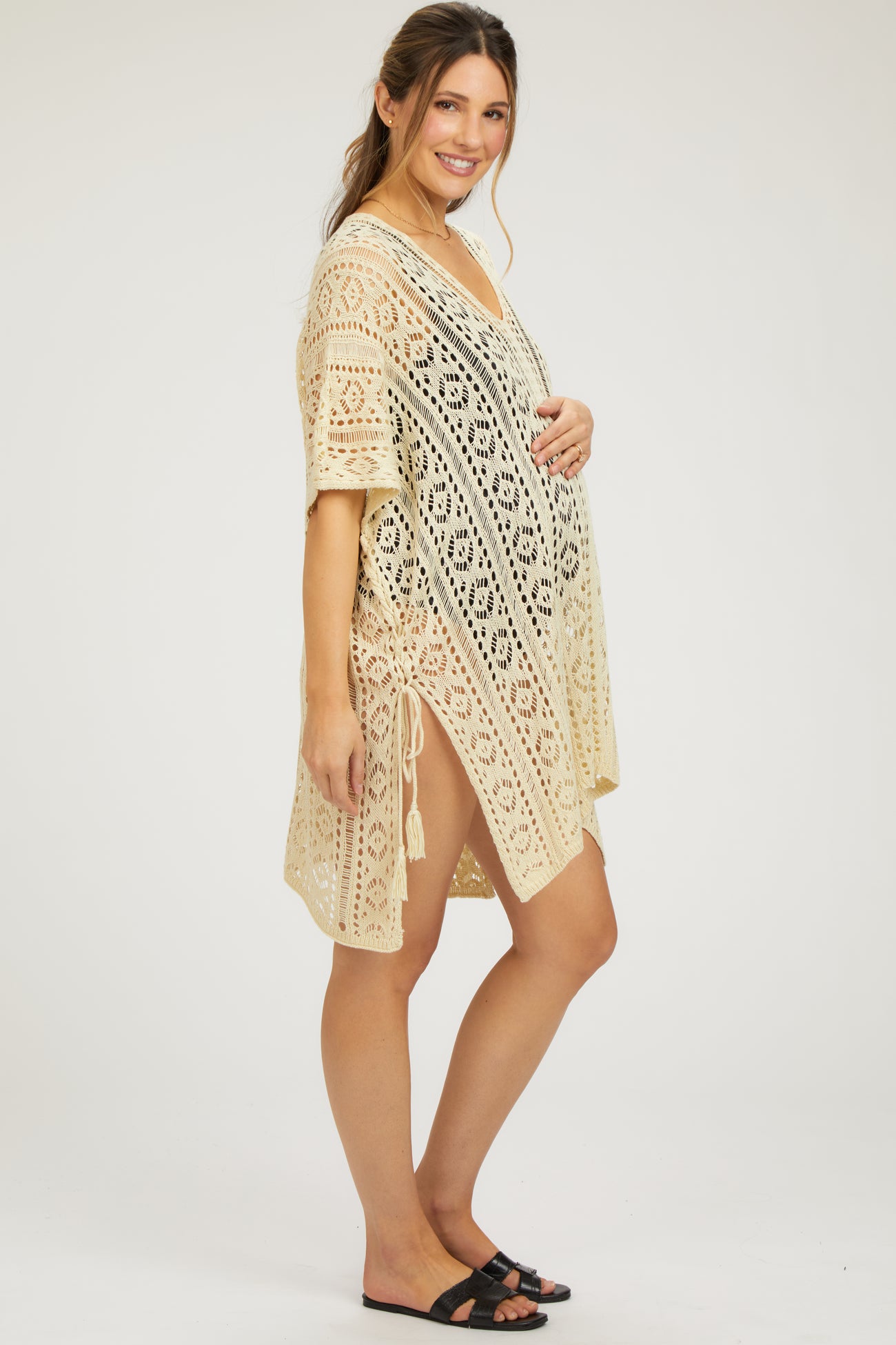woman wearing a beige crochet cover-up maternity wear with side ties and holding her baby bump