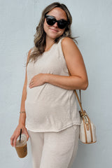 woman wearing a light beige sleeveless maternity top and beige pants, holding a drink and showing her baby bump