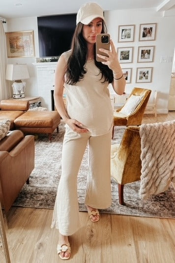 woman wearing a beige maternity tank top and matching flared pants, holding her baby bump while looking in the mirror