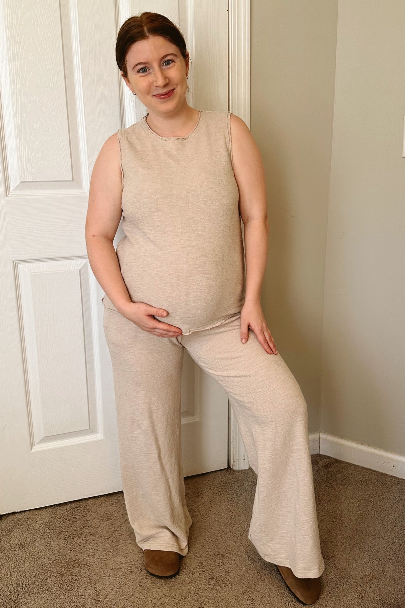 woman wearing a beige sleeveless maternity top and wide-leg pants, smiling and cradling her baby bump