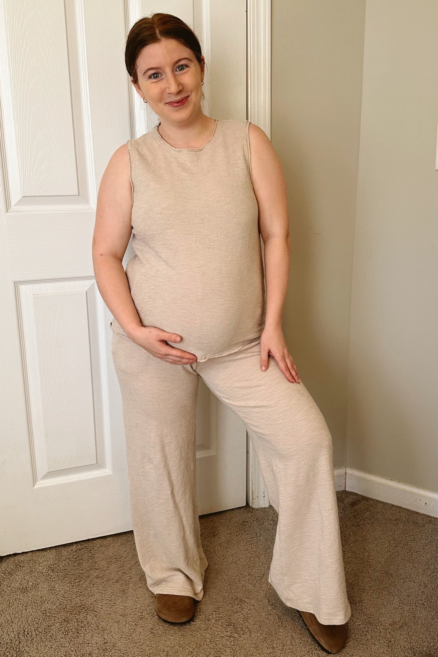 woman wearing a beige sleeveless maternity top and wide-leg pants, smiling and cradling her baby bump