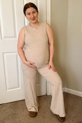woman wearing a beige sleeveless maternity top and wide-leg pants, smiling and cradling her baby bump