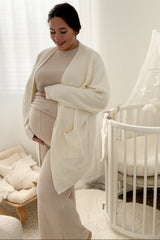 woman wearing a cream oversized cardigan over a beige maternity outfit with a visible baby bump, standing near a crib