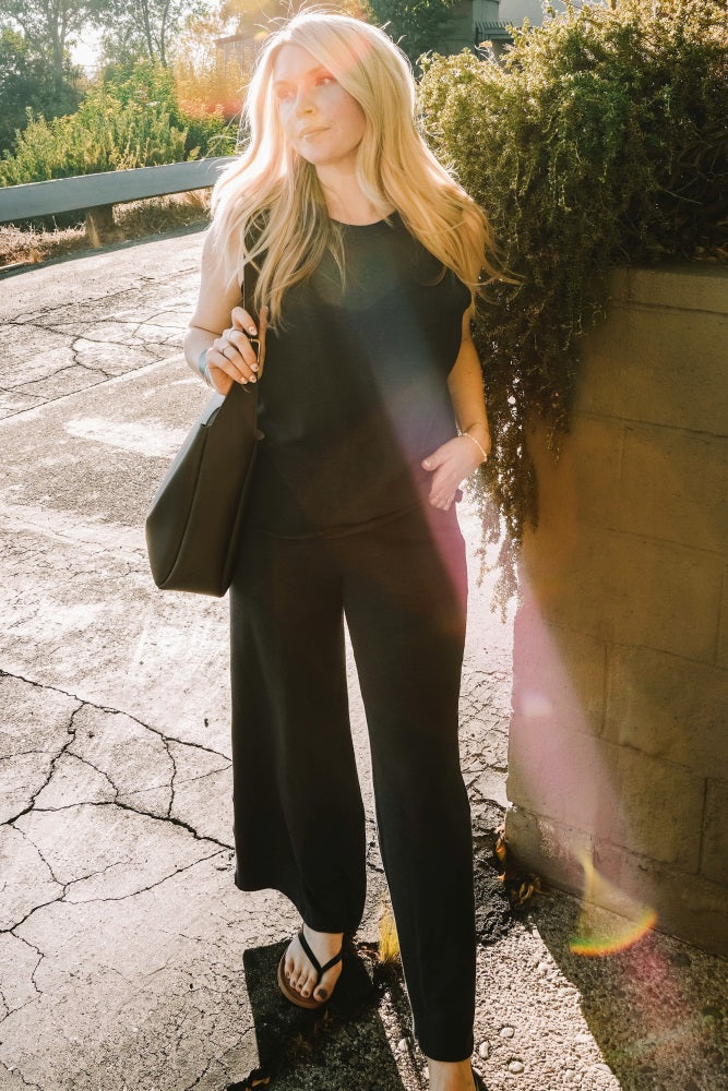 woman wearing a black sleeveless jumpsuit and carrying a black bag, standing outdoors with sunlight shining through trees