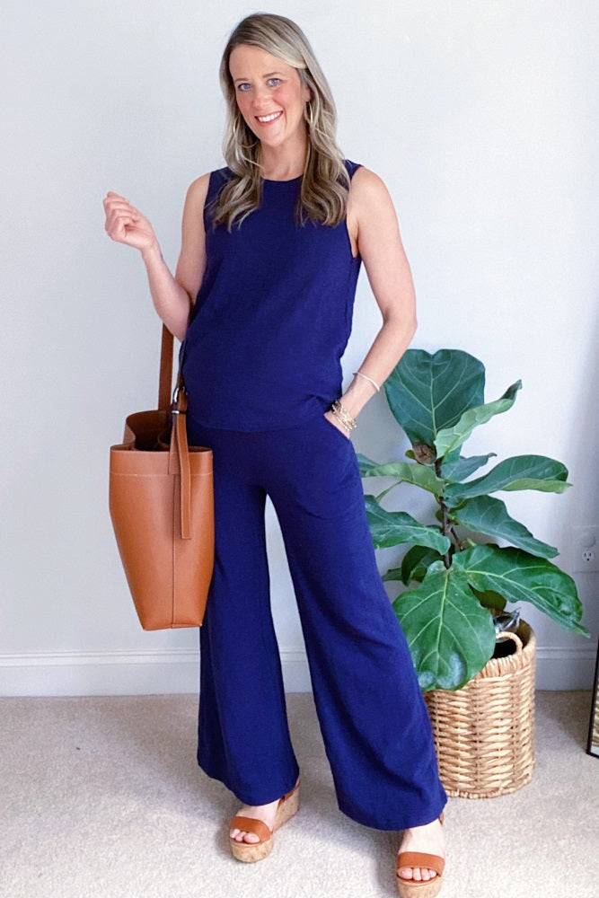 woman wearing a navy blue maternity tank top and wide-leg pants, posing with a tan tote bag and light brown wedges