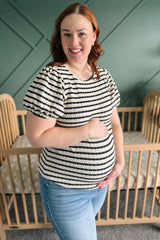 woman wearing a striped maternity top with puffed sleeves and holding her baby bump while standing near a crib