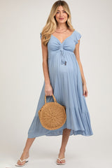 woman wearing a light blue maternity dress with tie front detail and flutter sleeves, holding a woven bag, and showcasing her baby bump