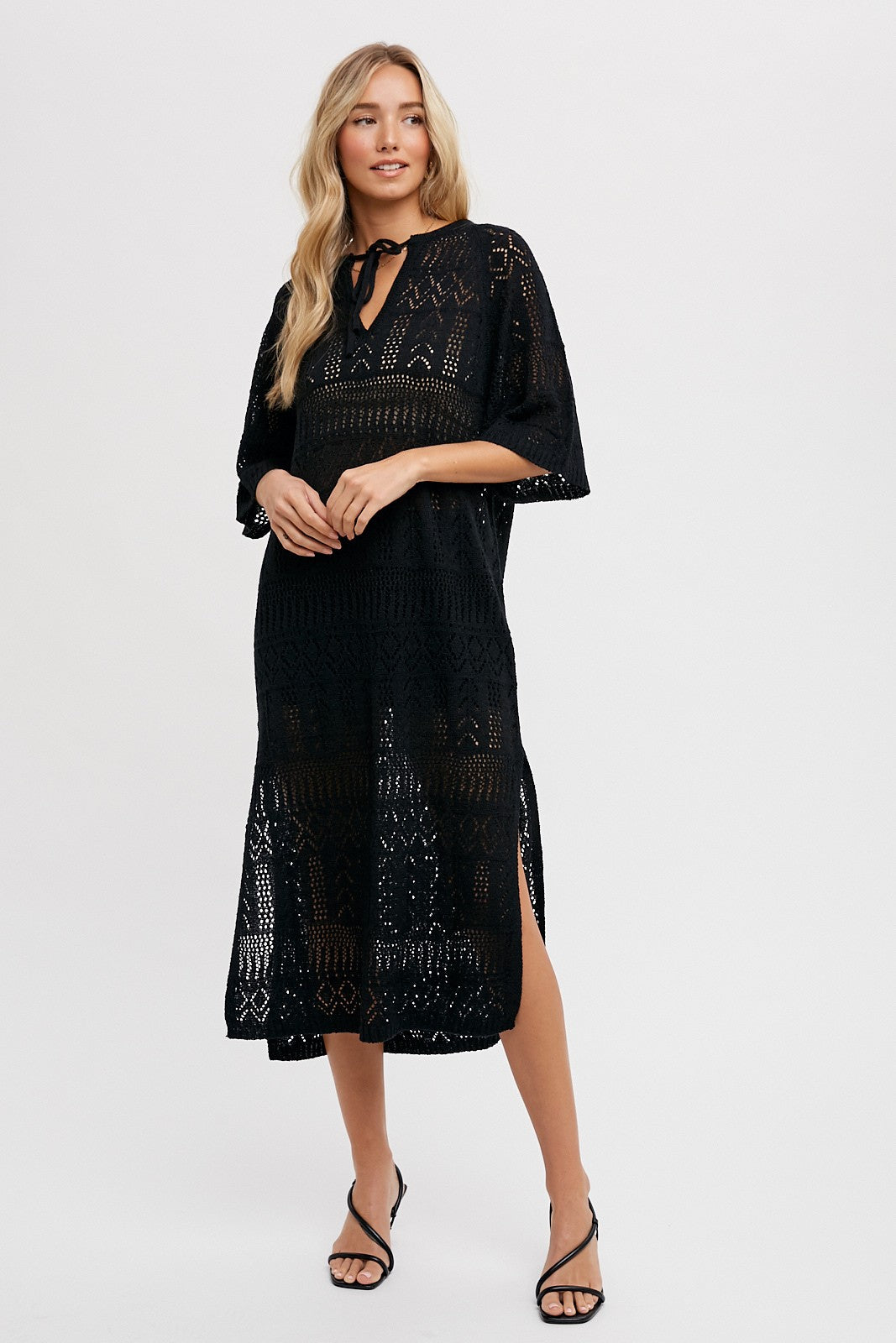 woman wearing a black crochet midi dress with a tie neckline and short sleeves, standing against a plain background