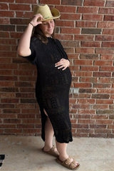 woman wearing a black maternity dress with side slits and a straw hat, showing her baby bump against a brick wall