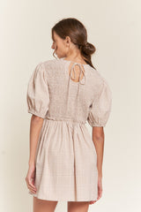 woman wearing a beige checkered smocked mini dress with puff sleeves and a tie closure at the back