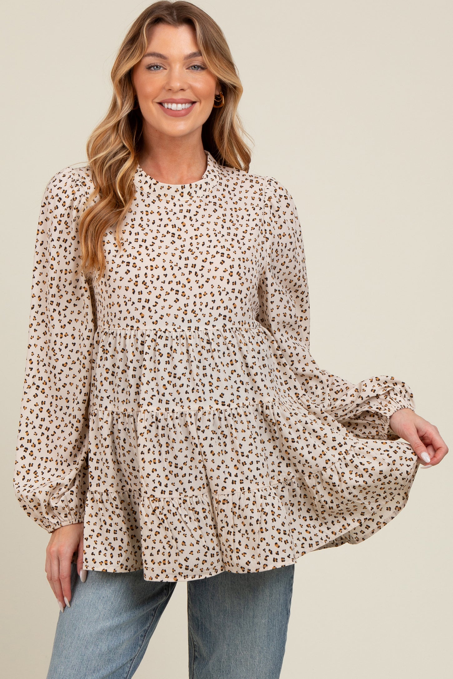 woman wearing a beige maternity top with a leopard print and tiered silhouette, smiling and holding the fabric out