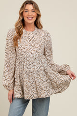 woman wearing a beige maternity top with a leopard print and tiered silhouette, smiling and holding the fabric out