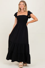 woman wearing a black maxi dress with ruffled sleeves and a smocked bodice, posing with one hand on her hip