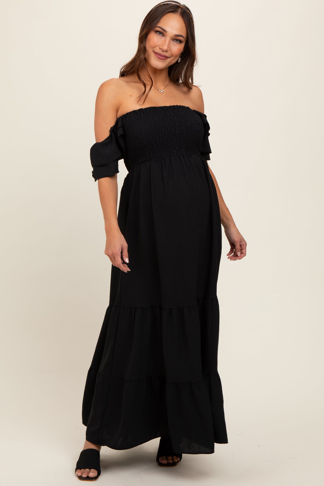 woman wearing a black off-the-shoulder maternity maxi dress with ruffled sleeves and a tiered skirt, showing her baby bump