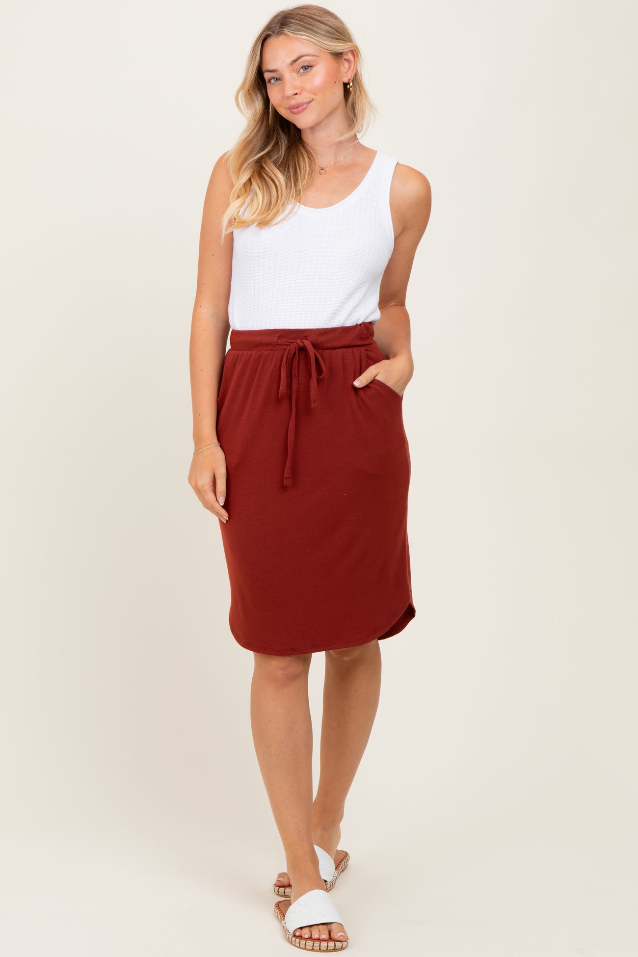 woman wearing a white ribbed tank top and a rust-colored drawstring skirt, standing in a relaxed pose