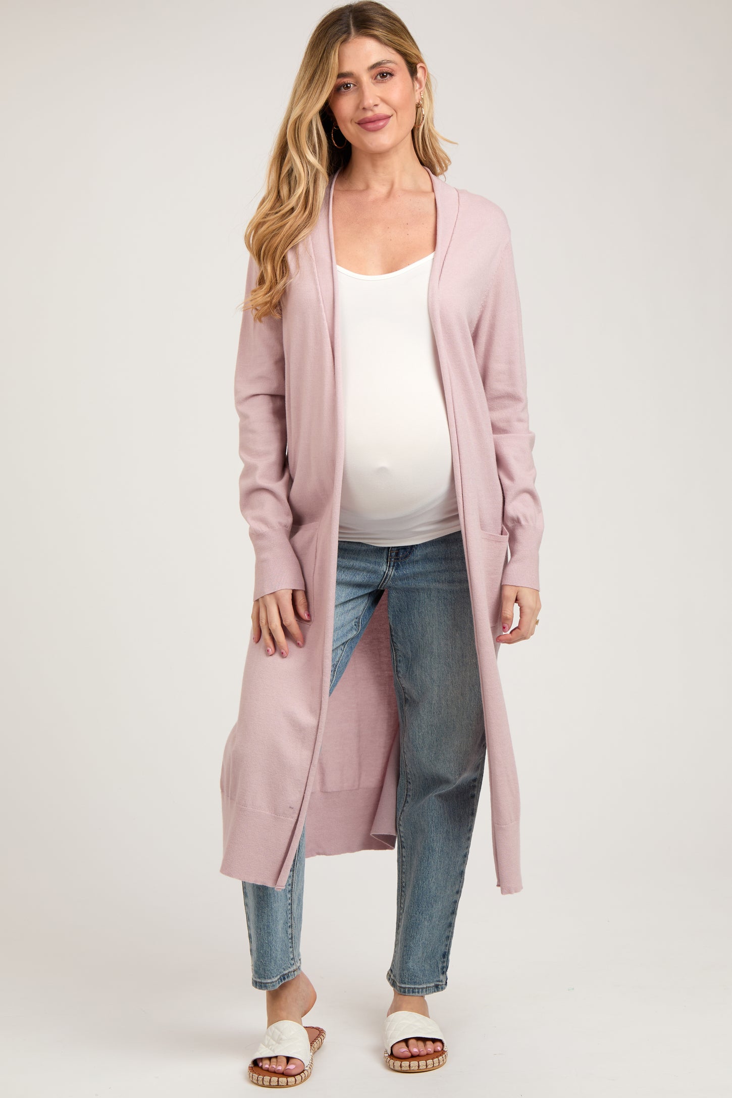 woman wearing a light pink long cardigan over a white tank top and blue jeans, showing her baby bump