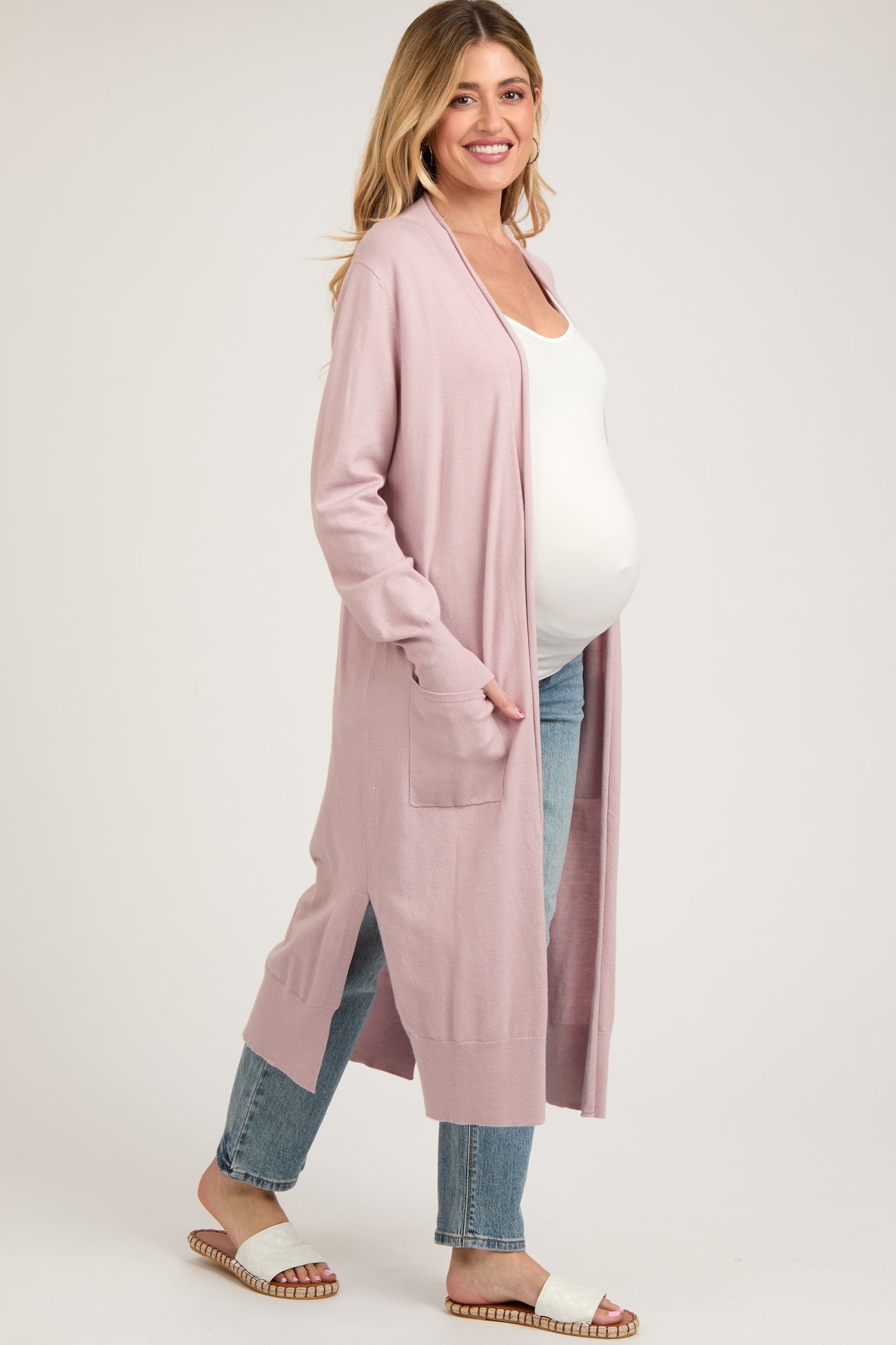 woman wearing a light pink maternity cardigan over a white tank top, with a visible baby bump, paired with light blue jeans