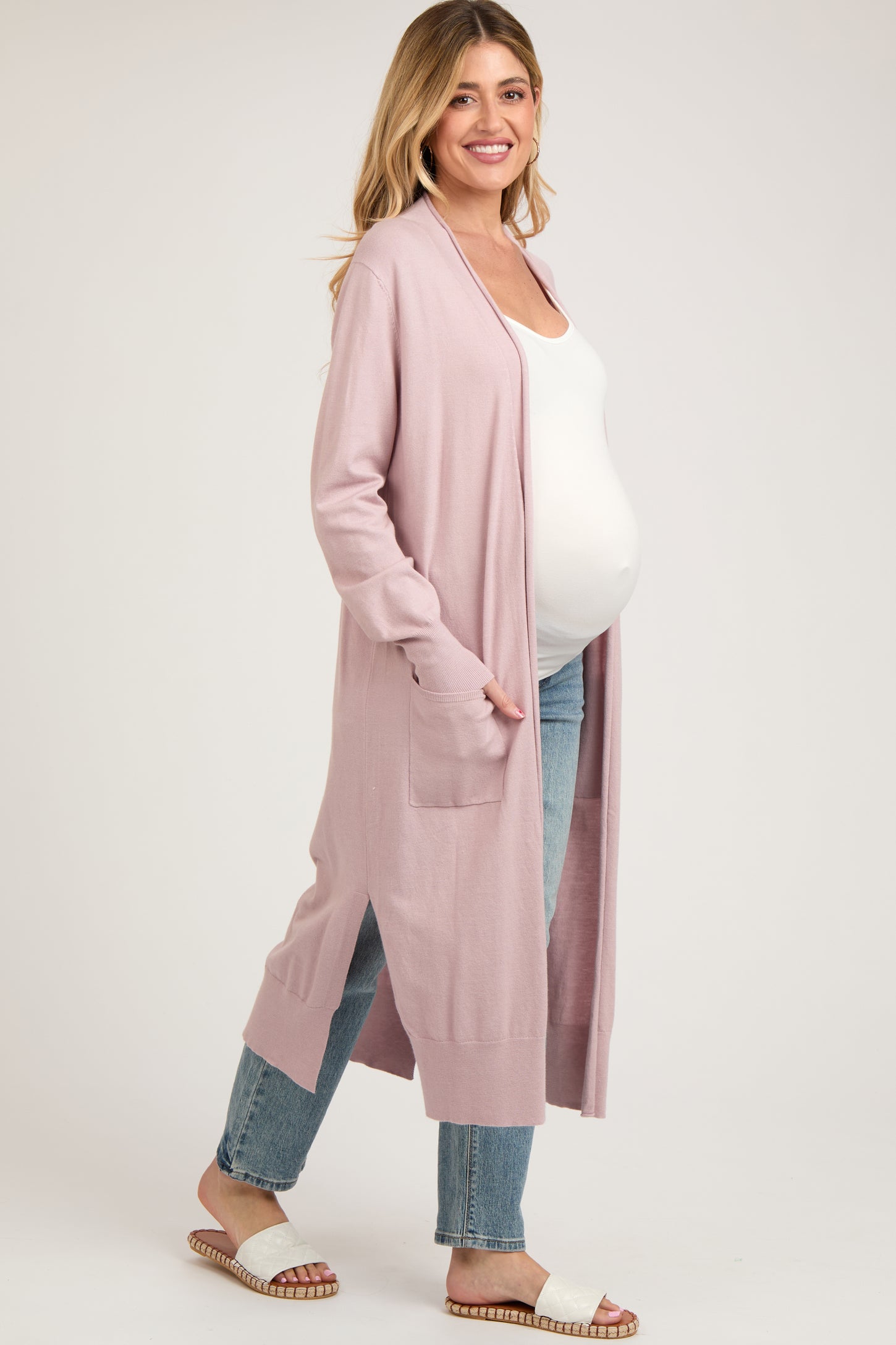 woman wearing a light pink maternity cardigan over a white tank top, with a visible baby bump, paired with light blue jeans
