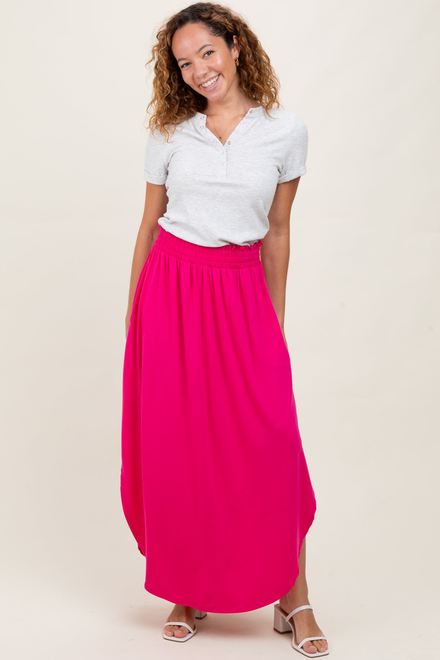 woman wearing a gray short sleeve henley top and a bright pink maxi skirt, standing and smiling