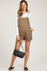 woman wearing a khaki maternity romper with adjustable straps and pockets, holding a black crossbody bag