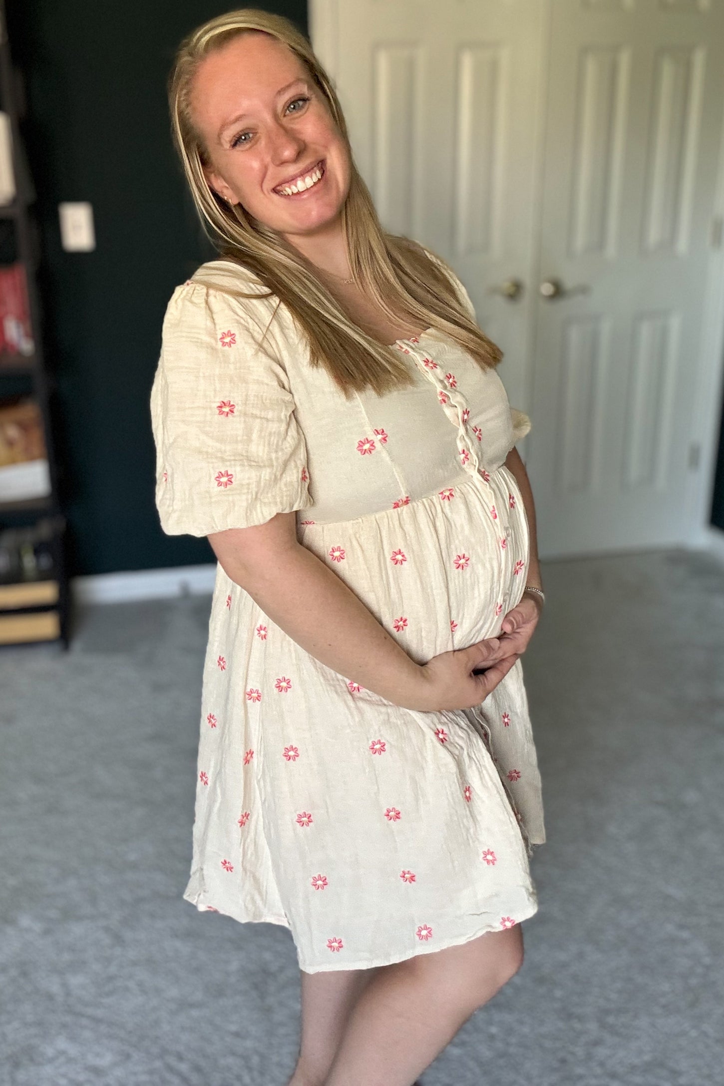 woman wearing a cream maternity dress with pink flowers and puff sleeves, showing her baby bump while smiling