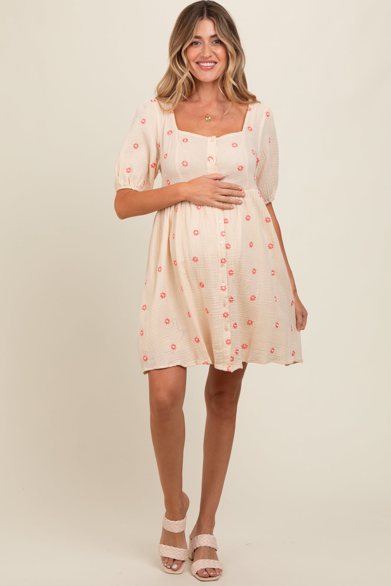 woman wearing a light beige maternity dress with pink floral prints and puff sleeves, holding her baby bump
