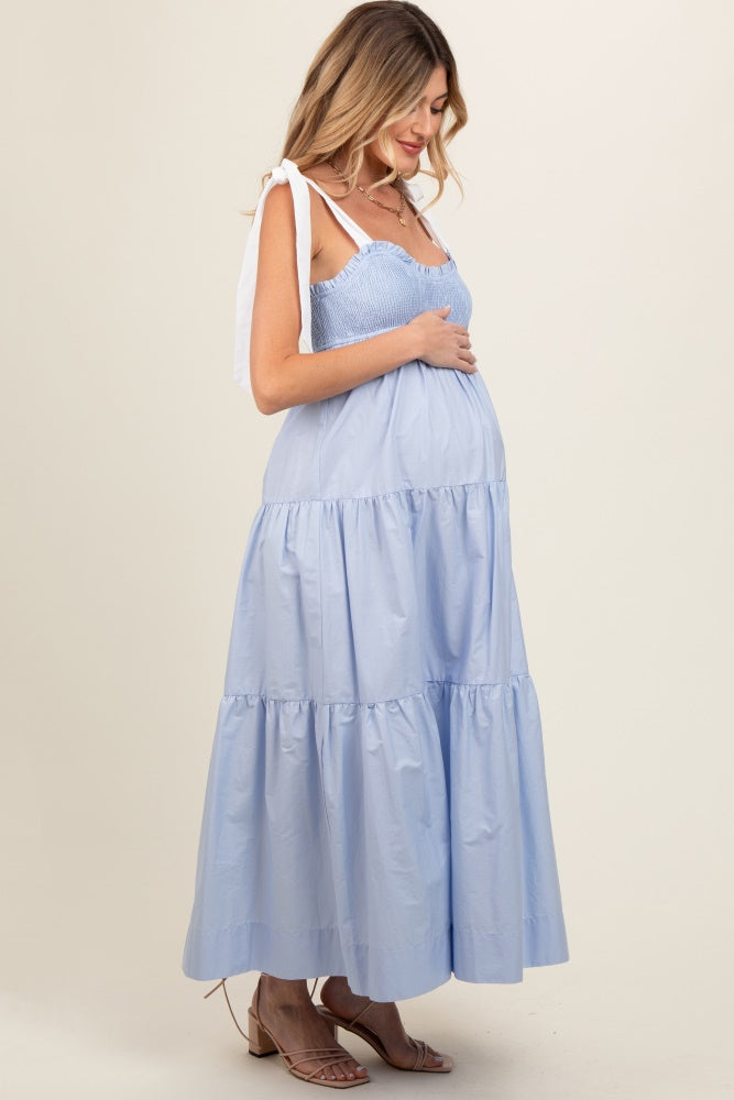 woman wearing a light blue maternity dress with tiered skirt and shoulder ties, gently holding her baby bump