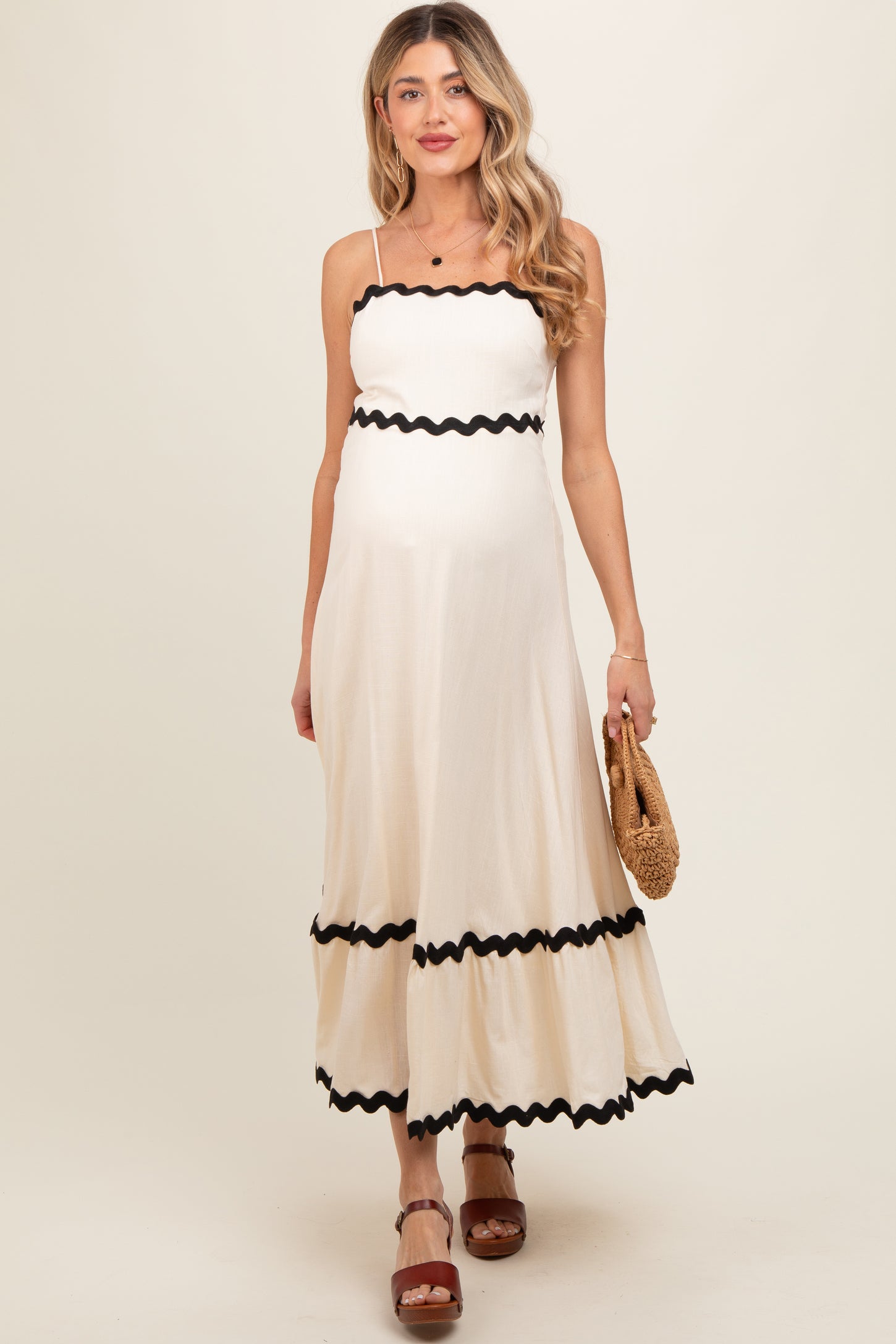 woman wearing a cream maternity maxi dress with black scalloped trim and holding a straw bag, showing her baby bump