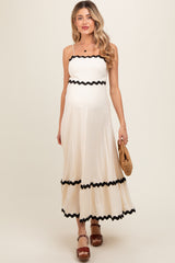 woman wearing a cream maternity maxi dress with black scalloped trim and holding a straw bag, showing her baby bump