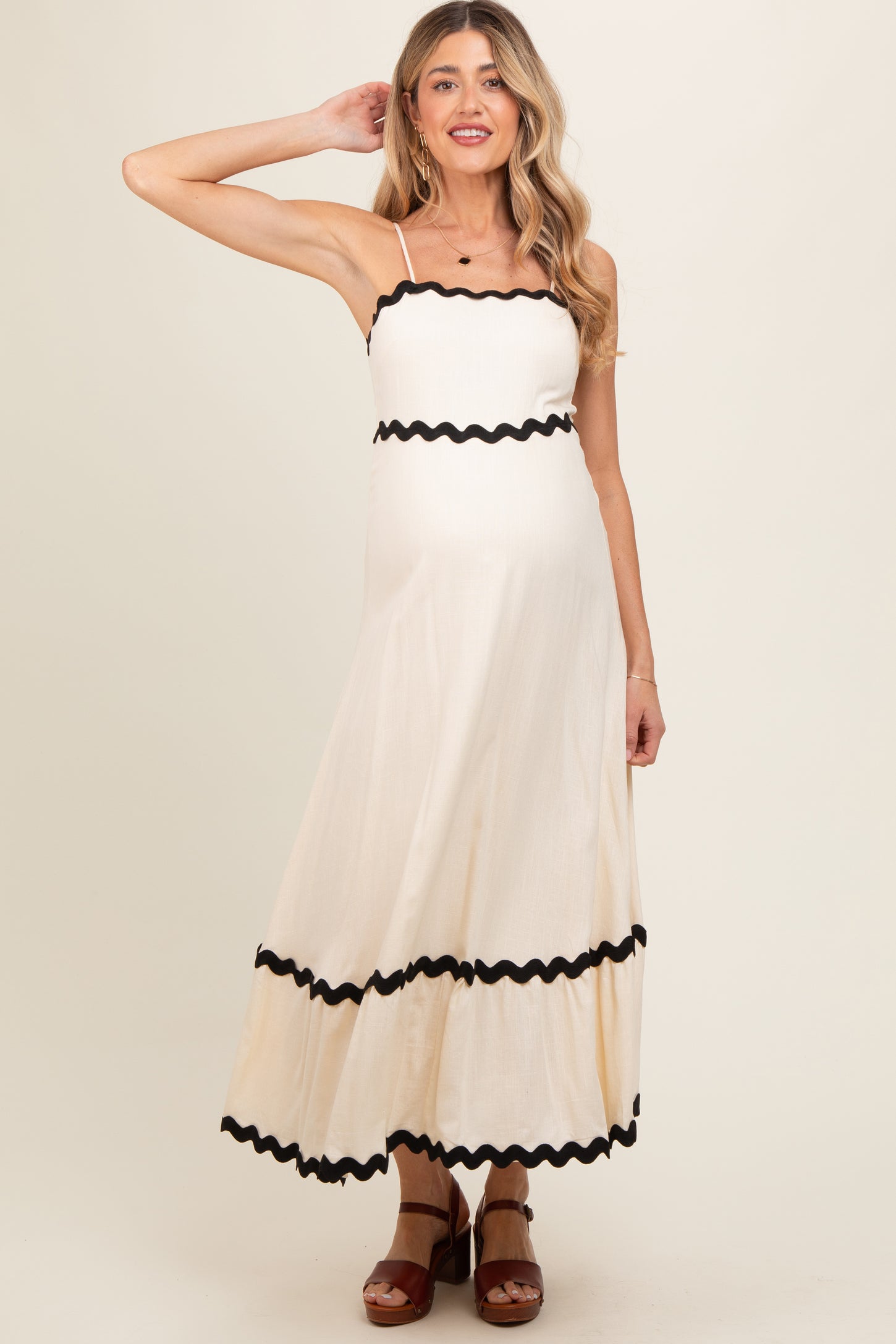 woman wearing a cream maternity maxi dress with black scalloped trim and spaghetti straps, showcasing her baby bump