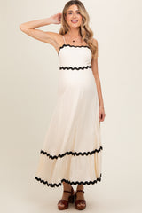 woman wearing a cream maternity maxi dress with black scalloped trim and spaghetti straps, showcasing her baby bump
