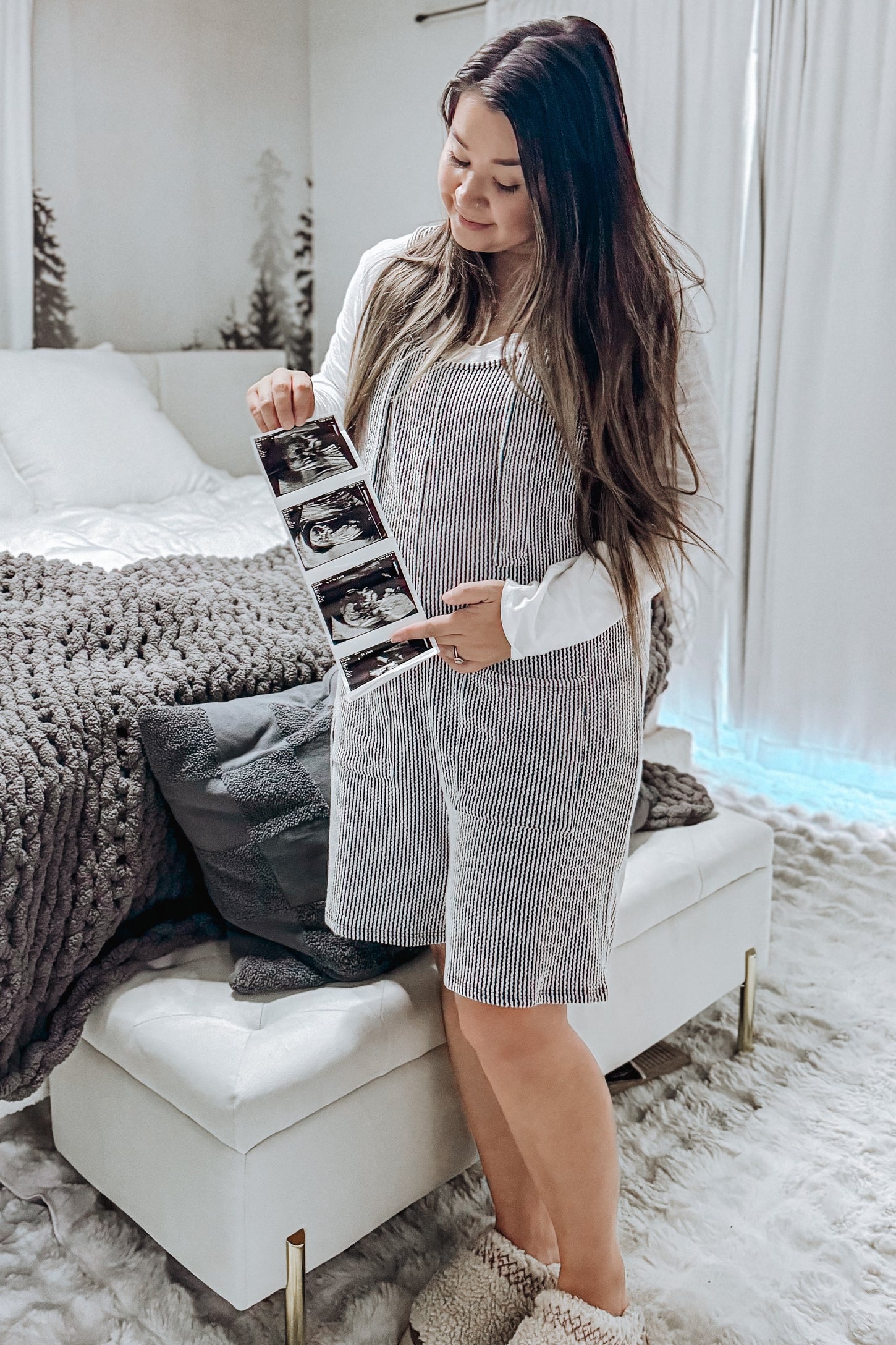 woman wearing a striped maternity dress holding ultrasound photos and looking at them while standing in a cozy bedroom setting