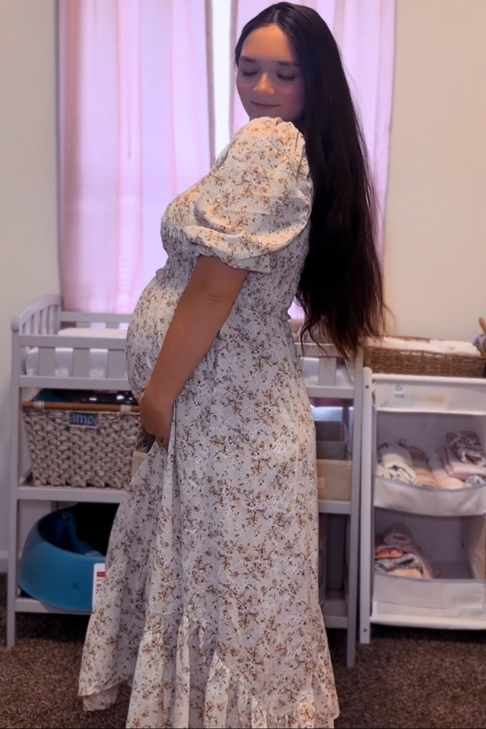 woman wearing a floral maternity dress, gently holding her baby bump with a soft smile in a cozy nursery setting