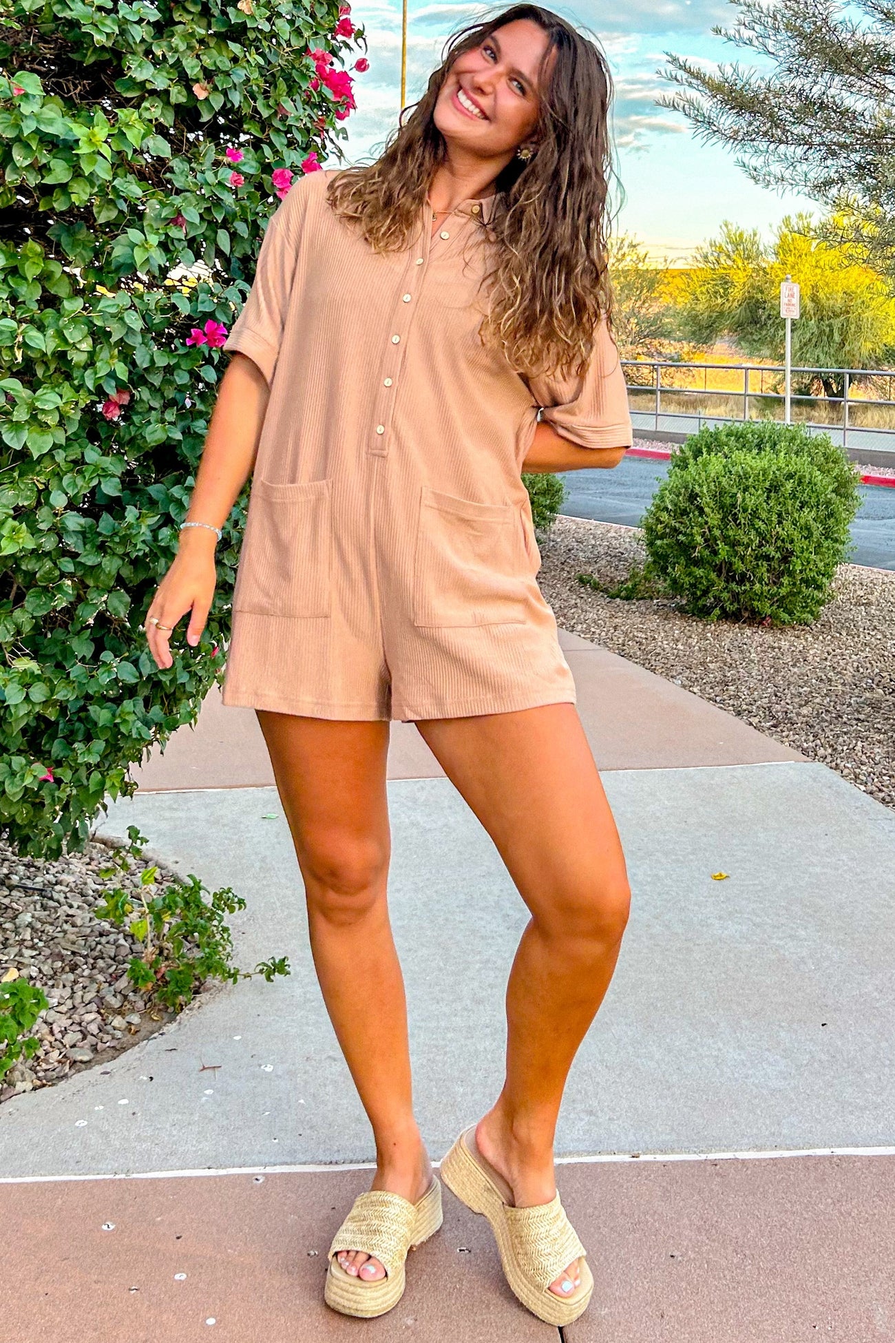 woman wearing a tan short sleeve romper with buttons and pockets, paired with wedge sandals and standing outside