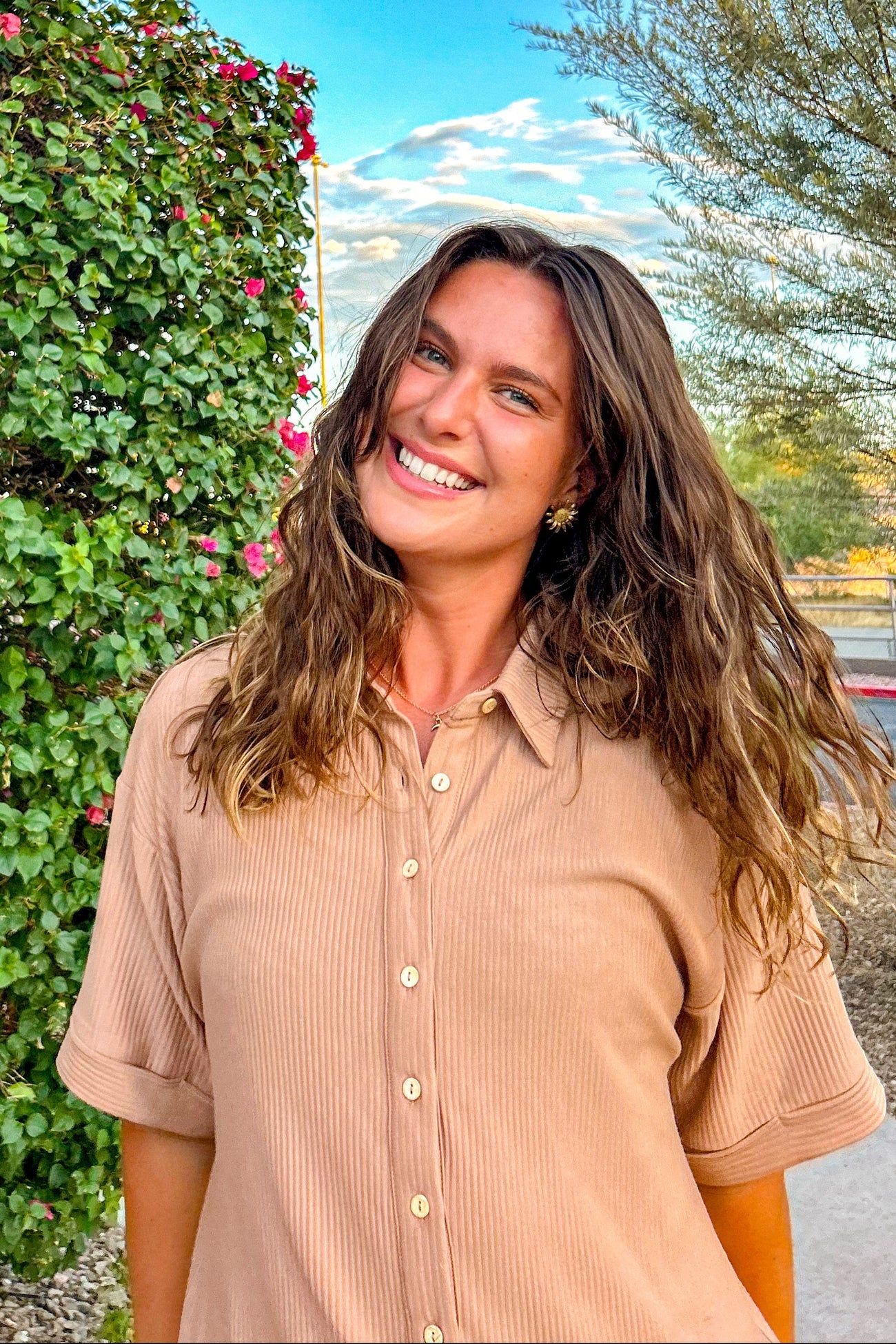 woman wearing a beige short sleeve button-up shirt, smiling outdoors with greenery and flowers in the background