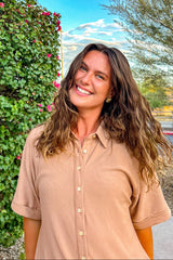 woman wearing a beige short sleeve button-up shirt, smiling outdoors with greenery and flowers in the background