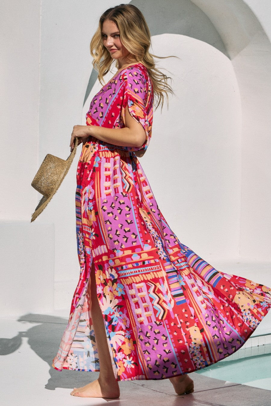 woman wearing a colorful maxi dress with a floral and geometric print, holding a straw hat, standing outdoors