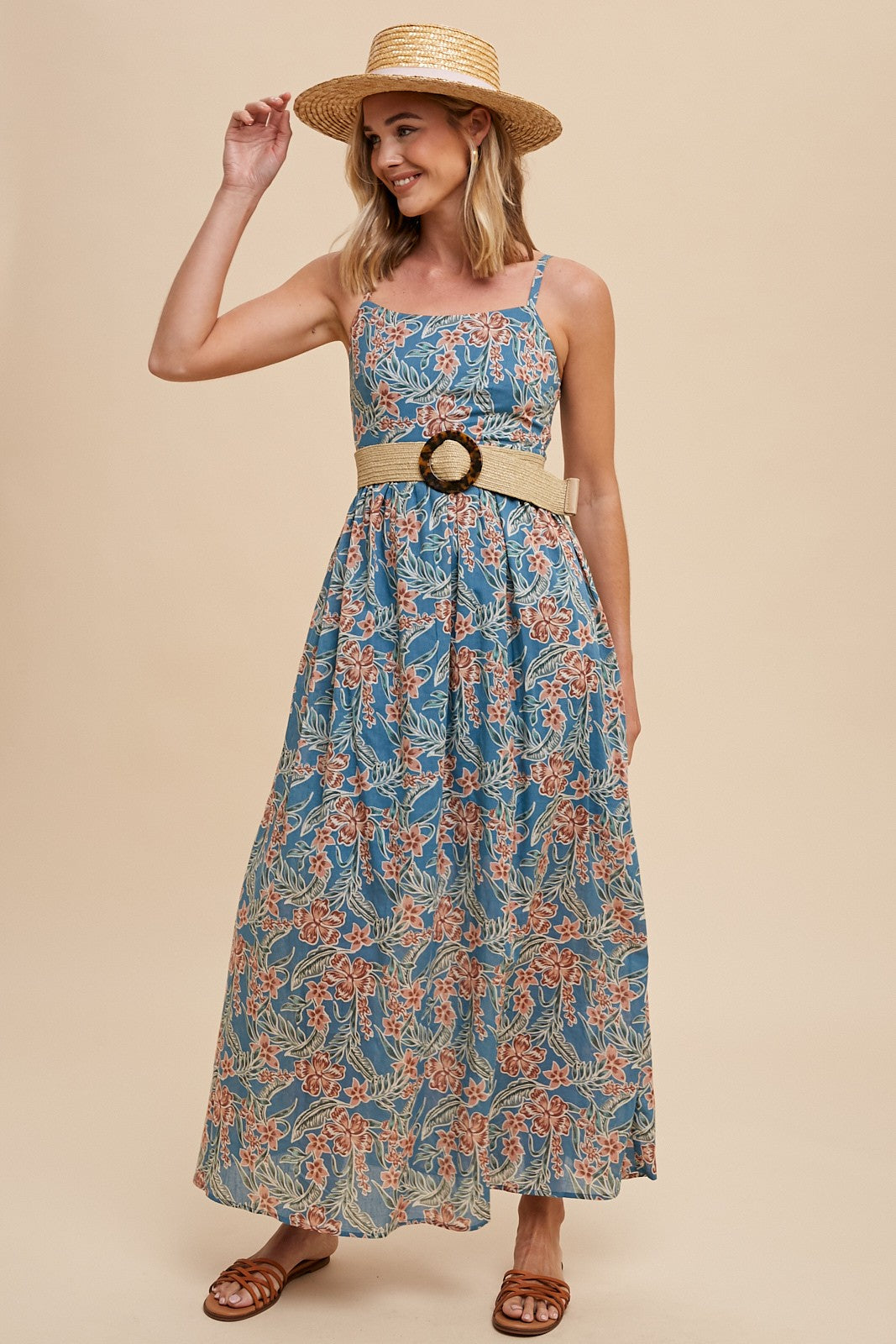woman wearing a blue floral maxi dress with a belted waist and a straw hat, smiling and posing with one hand raised