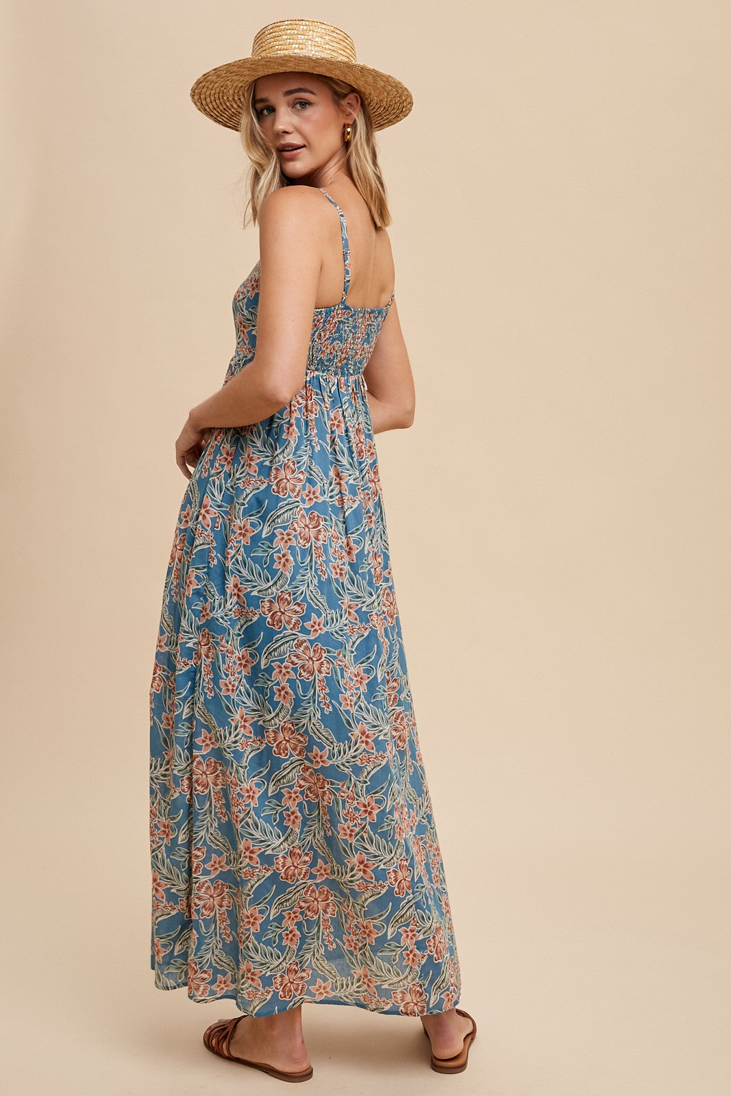 woman wearing a blue floral maxi dress with spaghetti straps, standing with a straw hat against a beige background