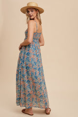 woman wearing a blue floral maxi dress with spaghetti straps, standing with a straw hat against a beige background