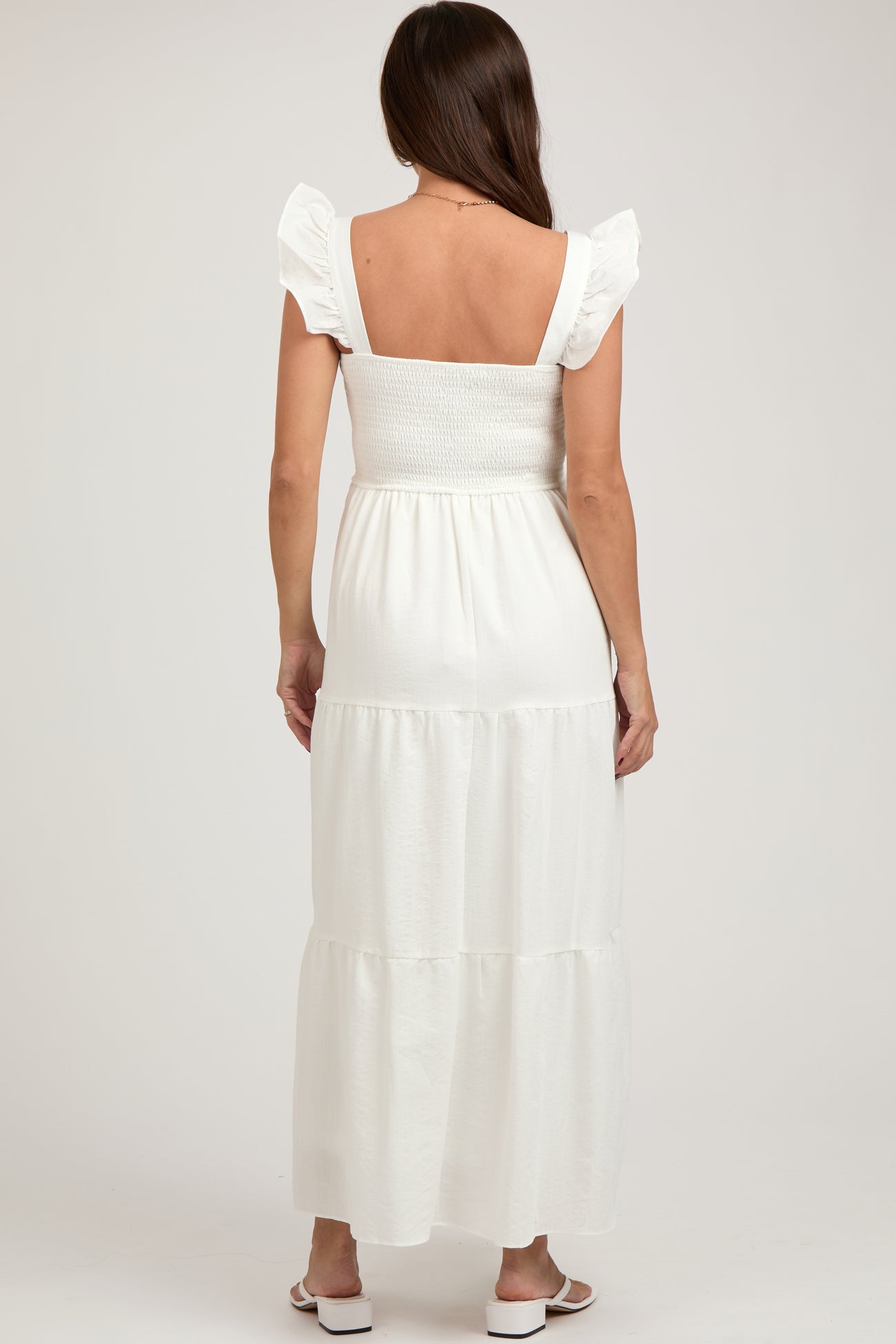 woman wearing a white maternity maxi dress with ruffled sleeves and a tiered skirt, showing a baby bump from the back
